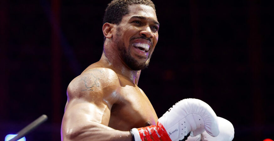 Boxer in the ring, smiling broadly while holding white boxing gloves and red hand wraps."