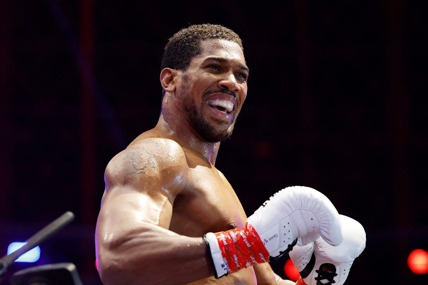 Boxer in the ring, smiling broadly while holding white boxing gloves and red hand wraps."